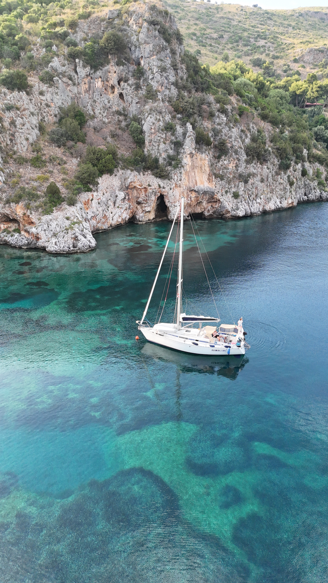 Aerial of Nene at anchor in a turquoise cove below cliffs